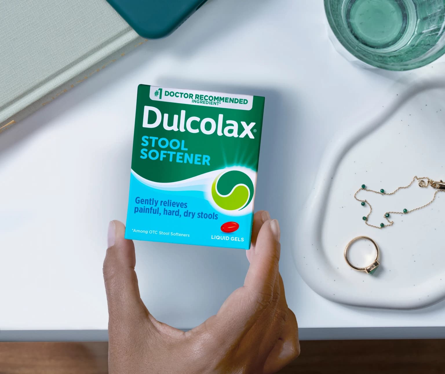 Hand holding Dulcolax Stool Softener box near a glass and jewelry.